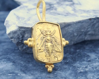 May include: Gold pendant featuring a detailed bee design, set in a rectangular frame with decorative accents. The pendant has a textured surface and a loop for attaching a chain.