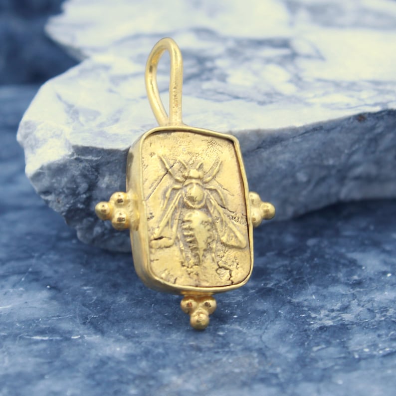 May include: Gold pendant featuring a detailed bee design, set in a rectangular frame with decorative accents. The pendant has a textured surface and a loop for attaching a chain.