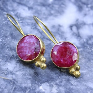 Puede incluir: Un par de pendientes dorados con grandes piedras preciosas redondas, facetadas, de color rojo rubí. Los pendientes tienen un diseño de gancho simple y un grupo de tres pequeñas cuentas doradas debajo de la gema.