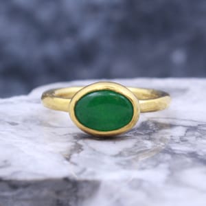 May include: A gold ring with a green oval gemstone. The ring is set on a white marble surface.