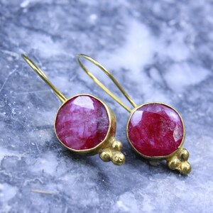 May include: A pair of gold-toned earrings featuring large, round, deep red gemstones. The gemstones are set in a gold bezel with decorative gold accents. The earrings have a simple hook design.