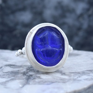 May include: A silver ring with a blue glass scarab beetle design. The ring has a beaded band.