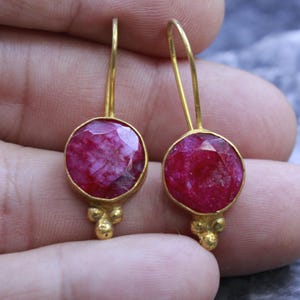 May include: A pair of gold-colored earrings with a round, faceted, ruby-colored stone. The earrings have a simple hook design and a decorative cluster of three small gold beads at the bottom of each stone setting.