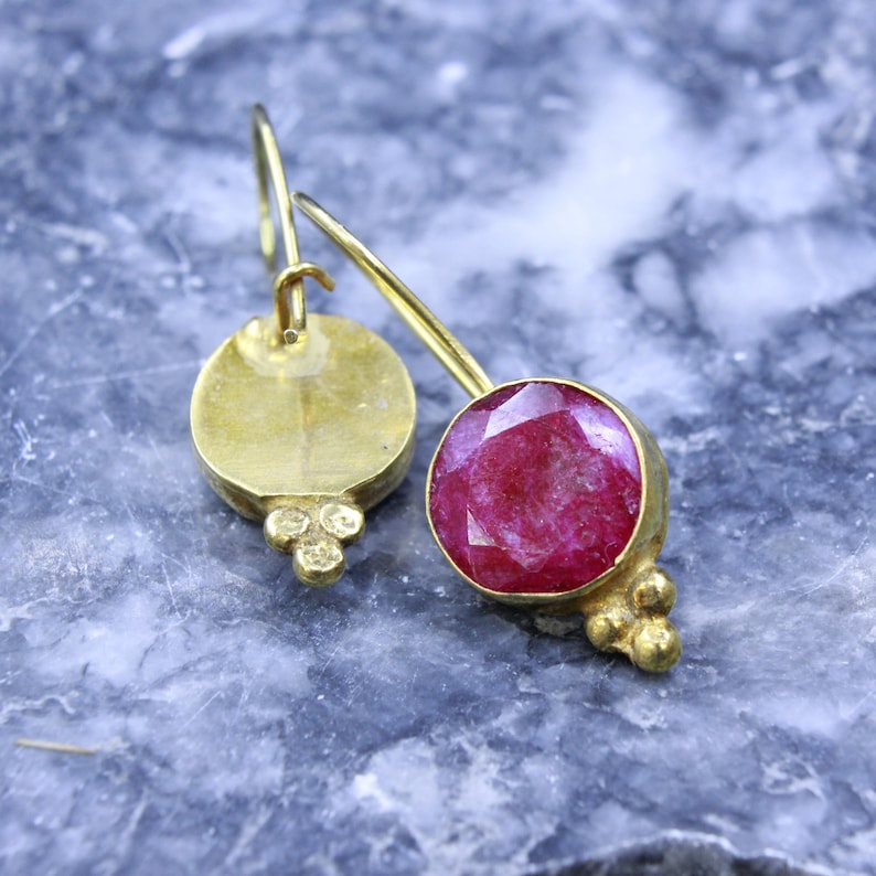 May include: A pair of gold-colored earrings with a round, faceted, ruby-colored gemstone. The earrings have a hook closure and a small cluster of gold-colored beads at the bottom of the gemstone setting. The background is a mottled blue and gray.