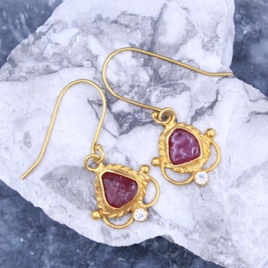 May include: Gold dangle earrings with a pink stone set in a gold frame and a small clear stone accent.