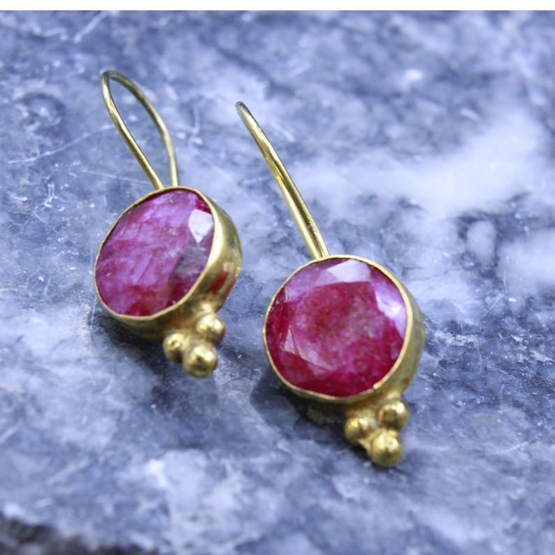 May include: A pair of gold-colored earrings with large, oval, ruby-red gemstones. The earrings have a simple hook design and small, decorative gold accents below the gemstones. The earrings are displayed on a textured, gray surface.
