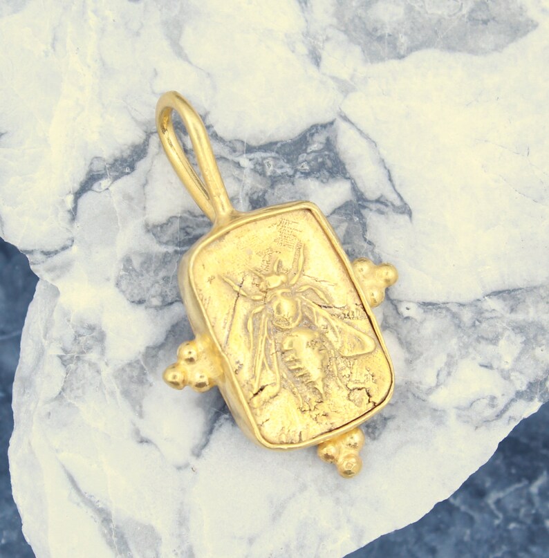 May include: Gold pendant featuring a detailed bee design. The pendant is rectangular with a textured surface and has a loop at the top for attaching to a chain.