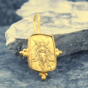 May include: Gold pendant featuring a detailed bee design. The pendant is rectangular with a textured surface and a loop at the top for attaching to a chain.