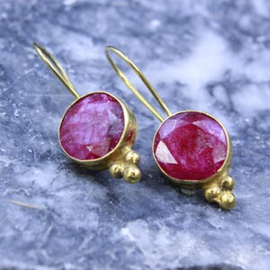May include: A pair of gold-colored drop earrings with large, round, faceted, ruby-red gemstones. The earrings have a simple, elegant design with a small cluster of gold beads at the bottom of each gemstone setting.