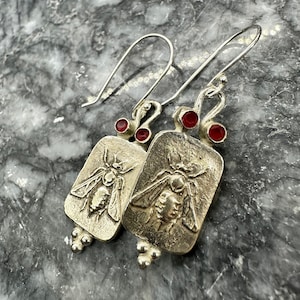 Puede incluir: Un par de pendientes de plata con un diseño de abeja y detalles de piedras preciosas rojas. Los pendientes son de forma rectangular y tienen una superficie texturizada.