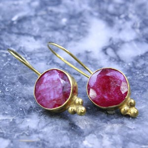May include: A pair of gold-colored earrings with large, round, faceted ruby gemstones. The earrings have a simple hook design and small, decorative gold accents. The ruby stones are a deep, vibrant red color.