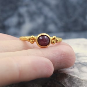 May include: A gold ring with a round, dark red gemstone set in the center. The ring has three small gold balls on each side of the stone.