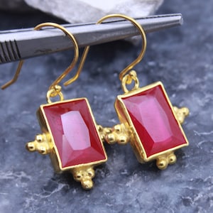May include: A pair of gold earrings with red gemstone rectangular drops. The earrings have a geometric design with small gold balls around the edges of the drops.