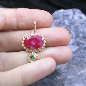 Puede incluir: Un colgante de oro con una piedra grande de forma ovalada en color rojo veteado, engastada en un marco de oro trenzado, con una piedra preciosa verde más pequeña y circular situada debajo, sujeta por una sencilla anilla de oro.