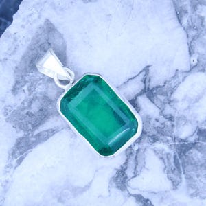 Puede incluir: Un colgante de piedra preciosa verde con corte esmeralda rectangular engastado en un bisel de metal plateado. El colgante presenta una superficie facetada y una anilla triangular en la parte superior, mostrando un color verde intenso.