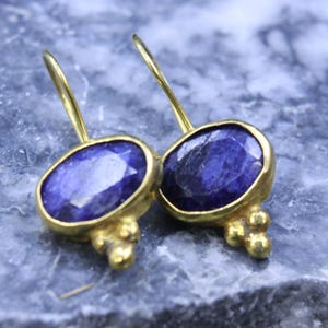 May include: A pair of gold-colored earrings featuring oval, deep blue gemstones. The gemstones are set in a gold bezel with a cluster of small gold beads at the base. The earrings have a simple hook design.