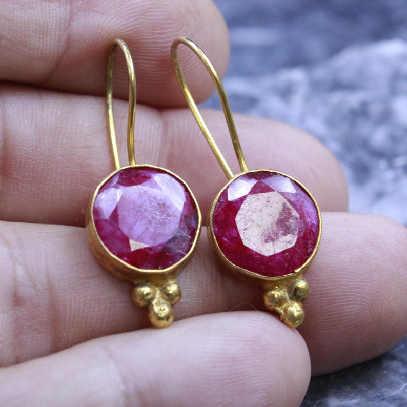 May include: A pair of gold-colored earrings featuring large, round, faceted ruby gemstones. The earrings have a simple design with a small cluster of gold beads at the bottom and a curved hook for the ear.