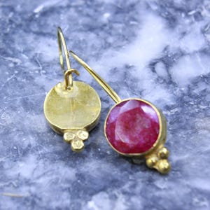 May include: A pair of gold-colored earrings. One earring features a round, faceted, deep red gemstone. The other earring has a gold disc with three small gold beads. Both earrings have a hook closure.