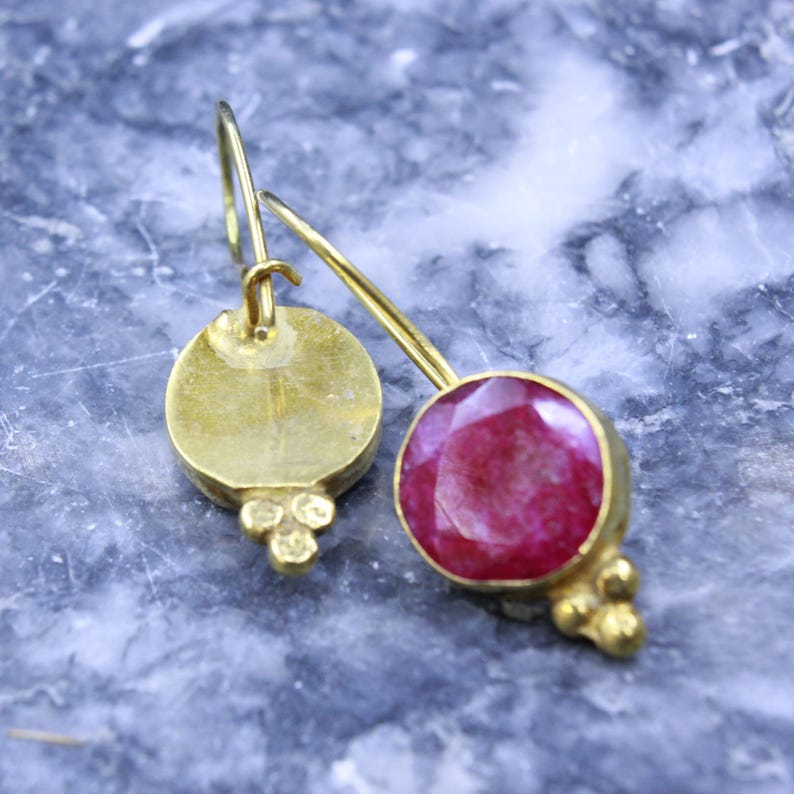 May include: A pair of gold-colored earrings. One earring features a round, faceted, deep red gemstone. The other earring has a gold disc with three small gold beads. Both earrings have a hook closure.