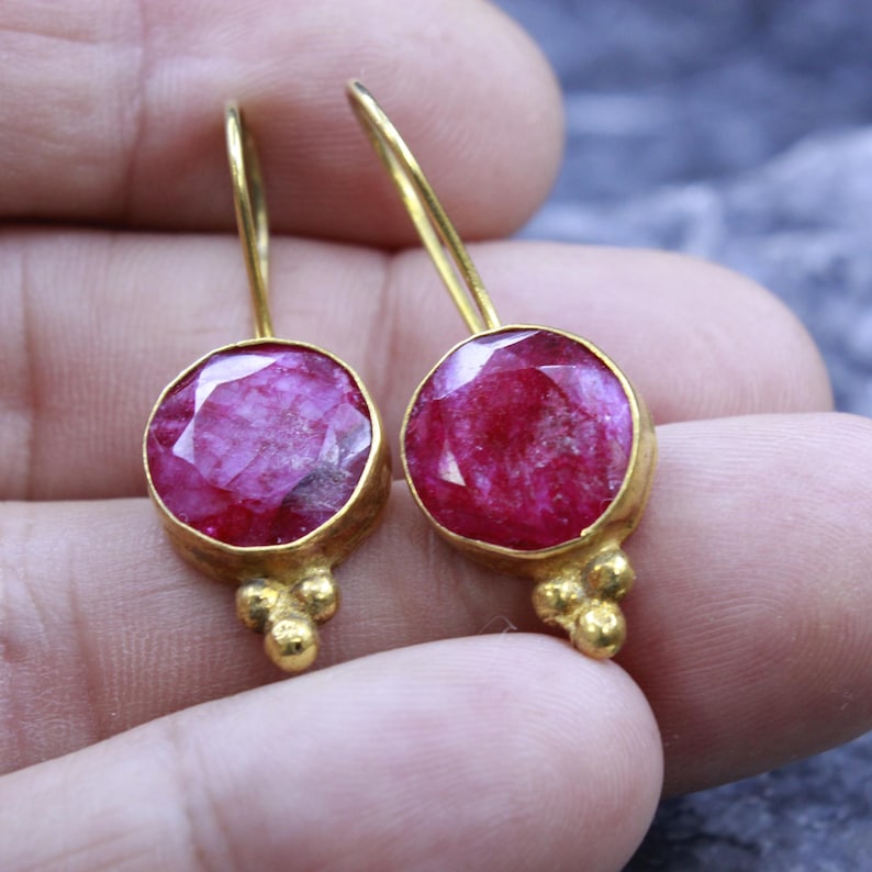 May include: A pair of gold-colored earrings featuring large, round, faceted, ruby-red gemstones. The earrings have a simple design with a small cluster of gold beads at the bottom of each earring.