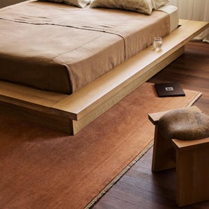 May include: A bedroom with a low wooden bed, brown bedding, and pillows. A matching rug and small stool with a cushion are in front of the bed. The room has a warm, neutral colour palette. The rug is approximately 200 cm in length.