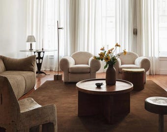 May include: A living room featuring a brown sofa, two cream-coloured armchairs, a round wooden coffee table with a dark vase of yellow flowers, a textured wooden chair, a small metal stool, and a dark brown area rug on a wooden floor.
