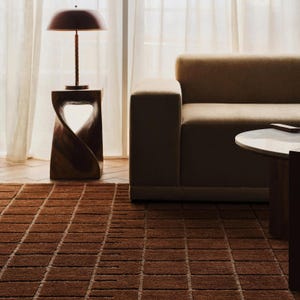 May include: A modern living room scene featuring a brown rug with a rectangular pattern, a brown sofa, and a wooden side table with a lamp. The lamp has a brown shade and a slender stem. The room has a neutral color palette.