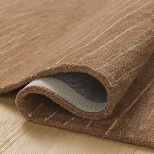 Op de afbeelding: Een opgerolde bruine vloerkleed met een getextureerd oppervlak en subtiele witte strepen. De onderkant van het vloerkleed onthult een grijze achterkant. Het vloerkleed ligt op een lichtgekleurde houten vloer.