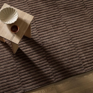 May include: A dark brown area rug with a repeating horizontal ribbed texture. A light-colored wooden side table sits on the rug, holding a white scalloped bowl and a small dark brown circular dish. The rug is placed on a light wood floor.