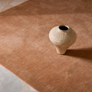 May include: A textured, light beige ceramic vase with a wide, rounded body and a narrow neck, featuring vertical carved grooves along its surface. The vase is positioned on a solid, warm-toned, rust-coloured rug with a soft, uniform pile texture.