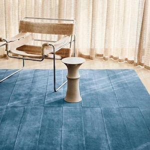 Könnte beinhalten: Ein modernes Interieur mit einem blauen Teppich mit strukturiertem, rechteckigem Muster. Ein beiger Stuhl mit Metallrahmen und ein kleiner, beiger Beistelltisch sind ebenfalls zu sehen. Transparente, beige Vorhänge hängen im Hintergrund und erzeugen ein weiches, diffuses Licht.