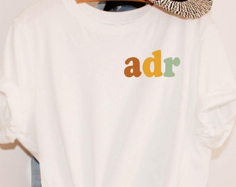 May include: White t-shirt with a colorful "adr" graphic on the front. The shirt is knotted at the bottom and is hanging on a hanger.
