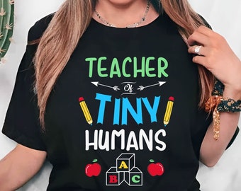 May include: A black t-shirt with the text "Teacher of Tiny Humans" in white and blue lettering. The shirt also features a red apple, a pencil, and colorful alphabet blocks.