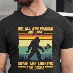 May include: A dark grey t-shirt with a vintage-style graphic of a Bigfoot holding a disc golf basket. The text reads "Not all who wander are lost. Some are looking for discs."