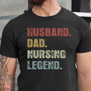 May include: A dark grey t-shirt with the text "Husband. Dad. Nursing Legend." printed in white with a vintage style font.