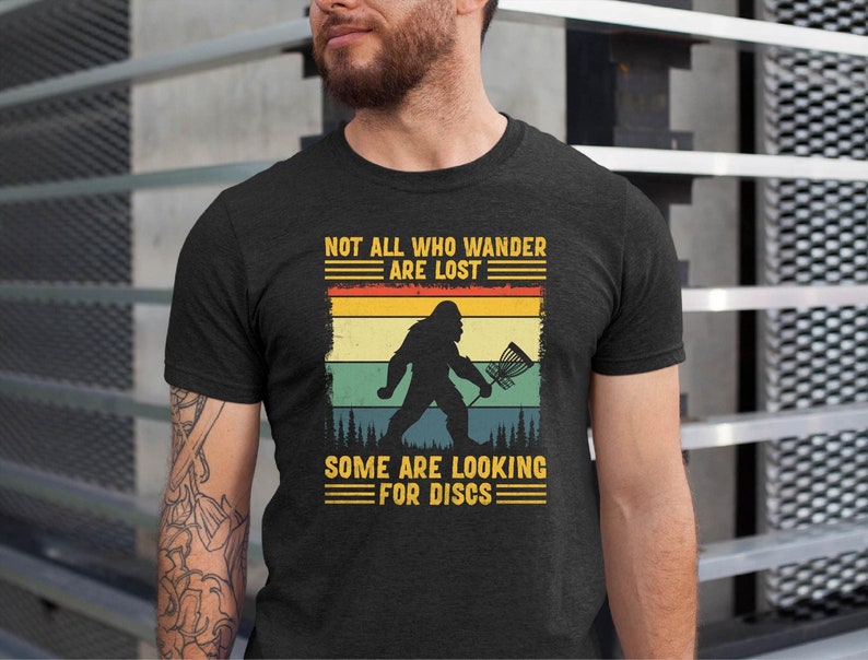 May include: A dark grey t-shirt with a vintage-style graphic of a Bigfoot holding a disc golf basket. The text reads "Not all who wander are lost. Some are looking for discs."