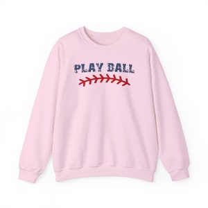 May include: Pink sweatshirt with the text "PLAY BALL" in blue with a red baseball stitch graphic.