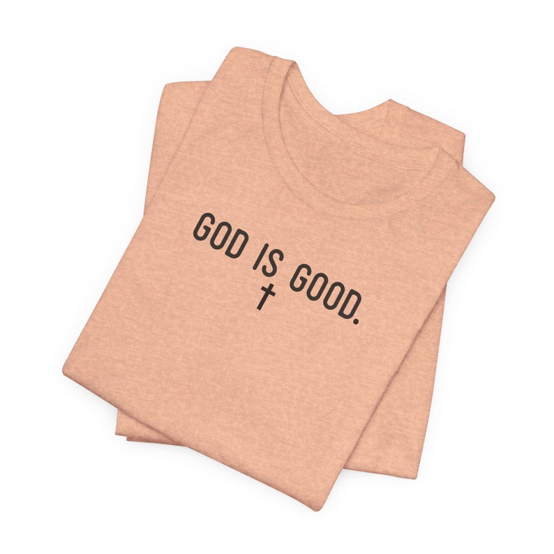 May include: A folded peach-colored t-shirt with the text "GOD IS GOOD." and a cross symbol printed on the front.