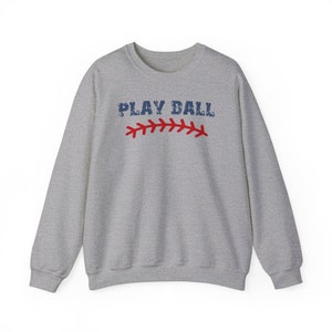 May include: A gray sweatshirt with the text "PLAY BALL" in blue and red stitching. The stitching is a baseball seam.