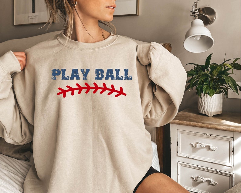 May include: A beige sweatshirt with the text "PLAY BALL" in blue with a red baseball stitch design.