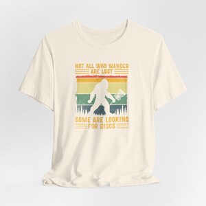 May include: A cream-colored t-shirt with a vintage-style graphic of a Bigfoot holding a frisbee. The graphic has a sunset background with the text "Not all who wander are lost. Some are looking for discs."