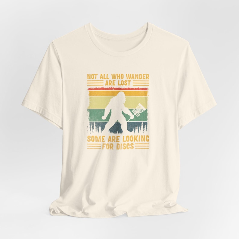 May include: A cream-colored t-shirt with a vintage-style graphic of a Bigfoot holding a frisbee. The graphic has a sunset background with the text "Not all who wander are lost. Some are looking for discs."