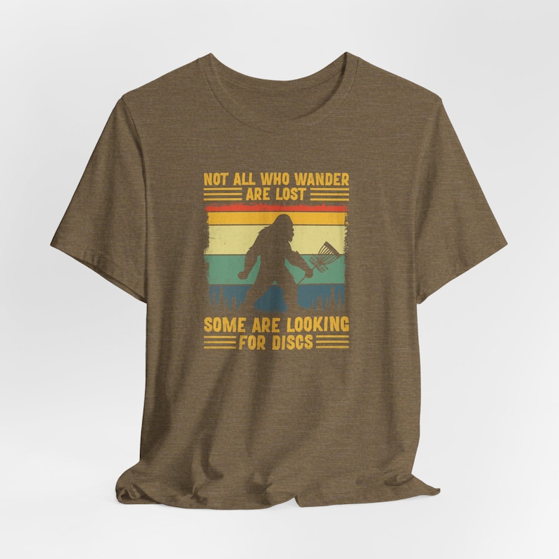 May include: A brown t-shirt with a vintage-style graphic of a Bigfoot holding a frisbee. The graphic says "Not all who wander are lost. Some are looking for discs."