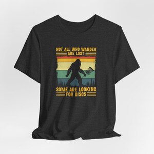 May include: A dark heather grey t-shirt with a vintage-style graphic of a Bigfoot holding a disc golf basket. The text "Not all who wander are lost. Some are looking for discs." is printed on the shirt.