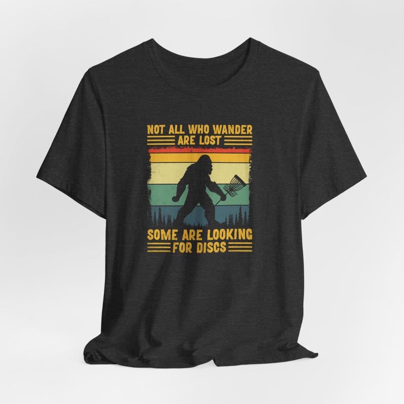 May include: A dark heather grey t-shirt with a vintage-style graphic of a Bigfoot holding a disc golf basket. The text "Not all who wander are lost. Some are looking for discs." is printed on the shirt.