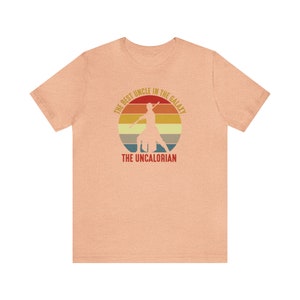 May include: A peach-colored t-shirt with a retro-style graphic of a person holding a lightsaber. The graphic says "The Best Uncle in the Galaxy, The Uncalorian".
