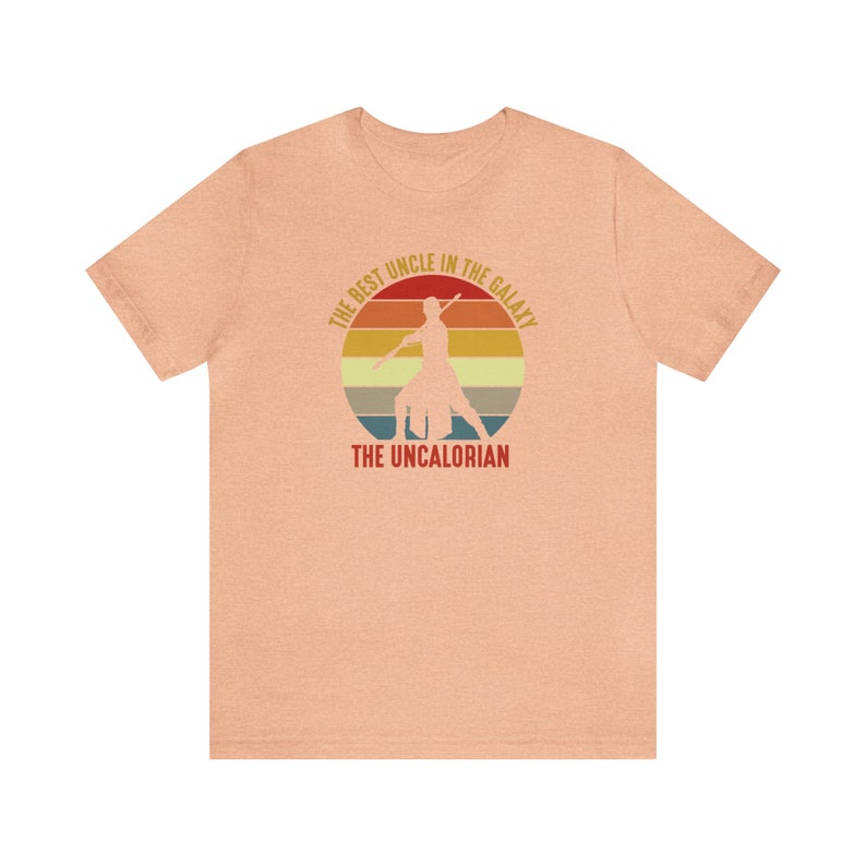 May include: A peach-colored t-shirt with a retro-style graphic of a person holding a lightsaber. The graphic says "The Best Uncle in the Galaxy, The Uncalorian".