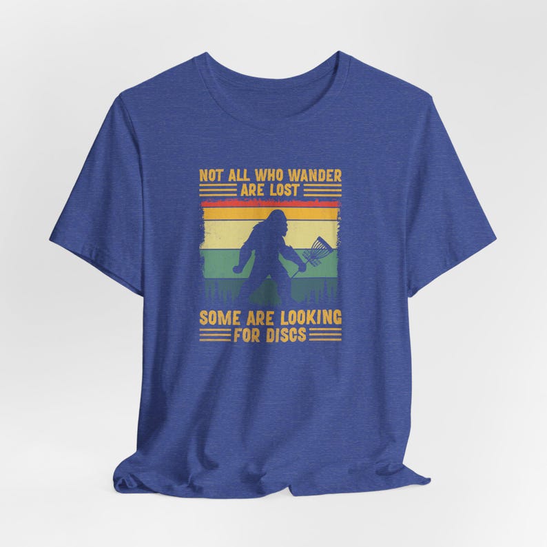 May include: A blue t-shirt with a vintage-style graphic of a Bigfoot holding a disc golf basket. The text reads "Not all who wander are lost. Some are looking for discs."