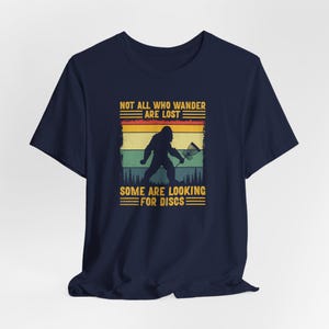 May include: A navy blue t-shirt with a vintage-style graphic of a Bigfoot holding a disc golf basket. The text reads "Not all who wander are lost. Some are looking for discs."