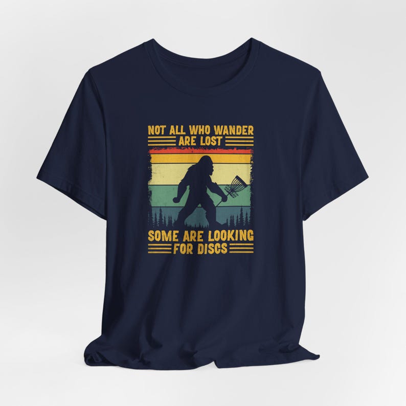 May include: A navy blue t-shirt with a vintage-style graphic of a Bigfoot holding a disc golf basket. The text reads "Not all who wander are lost. Some are looking for discs."